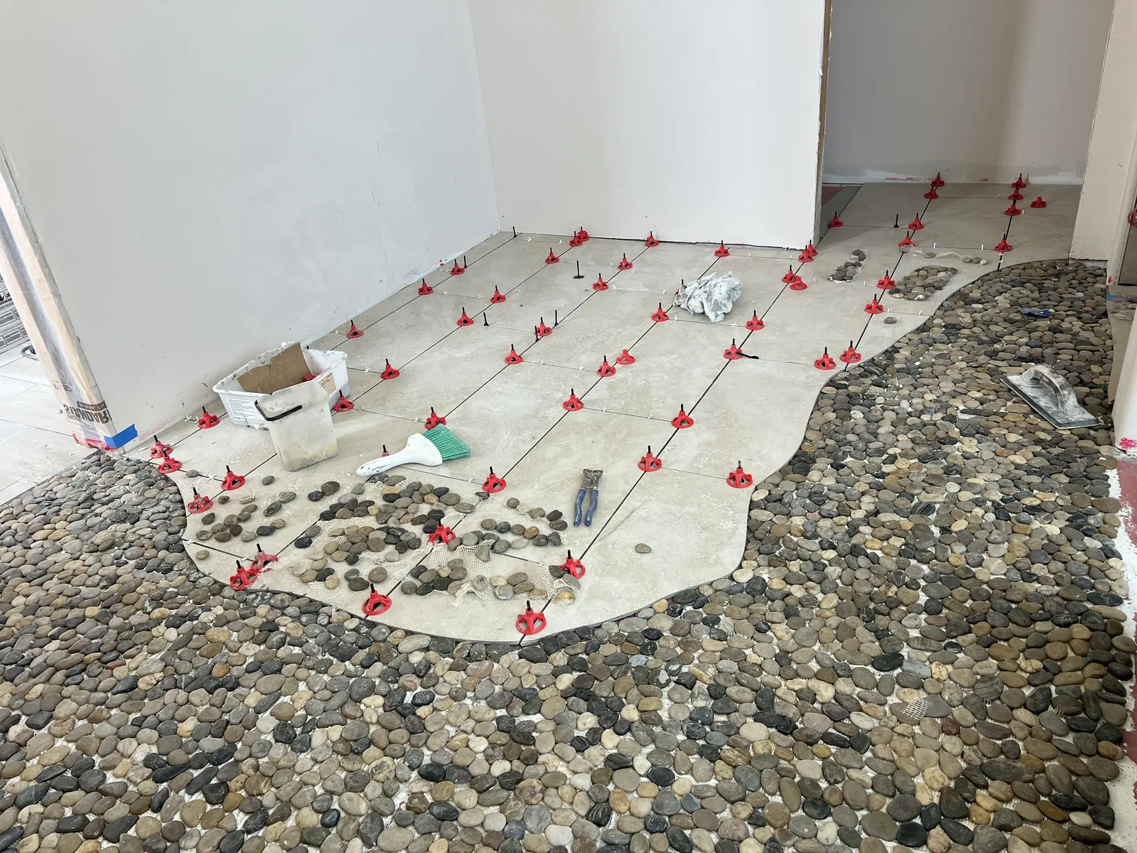 Travertine floor tile hand-cut to a flowing curve where it meets a river-rock floor, with leveling clips still in place