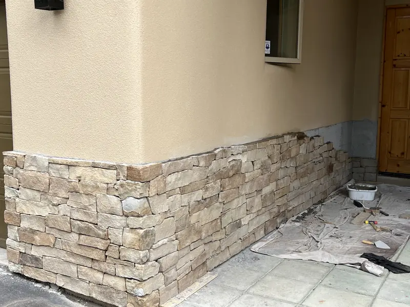 Stacked ledgestone veneer wainscot along a stucco wall beside a covered entry